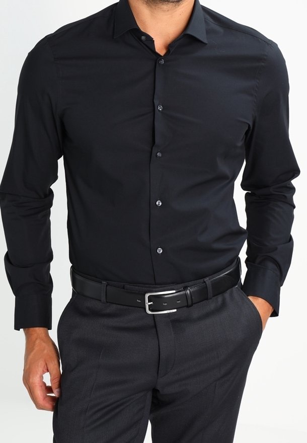 Homme portant une chemise noire ajustée à boutons, une ceinture en cuir noir à boucle argentée et un pantalon gris foncé, une main dans la poche.