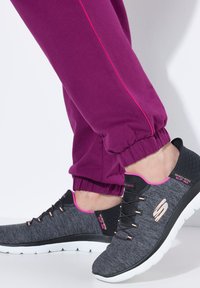 Schwarze und graue Sportschuhe mit rosa Akzenten, ausgestattet mit einem strukturierten Obermaterial aus Stoff, Schnürsenkeln und einer gepolsterten weißen Sohle, kombiniert mit lila Jogginghosen.