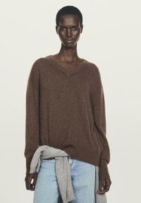 Pull en laine marron à col en V avec une coupe décontractée et des poignets côtelés, associé à un jean bleu clair et un article gris noué à la taille.