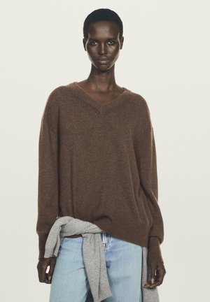 Maglione bordeaux a collo a V in maglia con vestibilità ampia e polsini a costine, abbinato a jeans blu chiaro e un capo grigio annodato in vita.