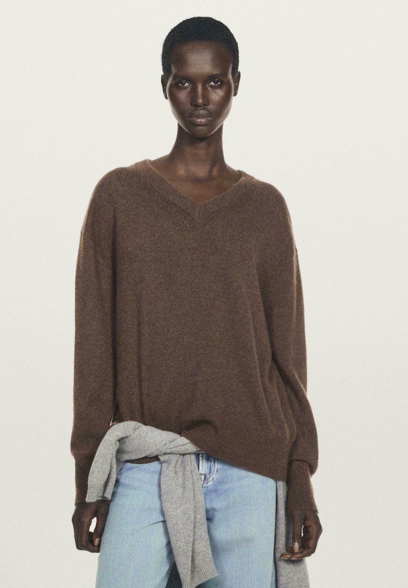 Pull en laine marron à col en V avec une coupe décontractée et des poignets côtelés, associé à un jean bleu clair et un article gris noué à la taille.