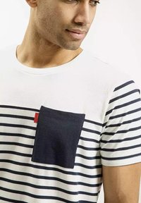 Pasiasta koszulka z krótkim rękawem w białym i granatowym kolorze, z jednolitym granatowym kieszonką i czerwoną metką. Miękka tkanina o luźnym kroju.