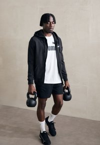 Sort zip-hoodie over en hvid t-shirt, sorte shorts og sportsko. Holder to sorte kettlebells med 12 kg mærkning. Ensartet baggrund.