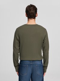 Oliwkowozielona koszula z długim rękawem wykonana z fakturowanej tkaniny, z okrągłym dekoltem, noszona z niebieskimi dżinsami, pokazująca gładki tył.