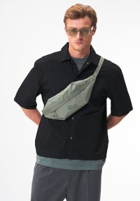 Helles grünes Stoff-Slingbag, quer über den Körper getragen, mit einem Reißverschluss und zwei runden Akzenten, kombiniert mit einem schwarzen Kurzarmhemd.