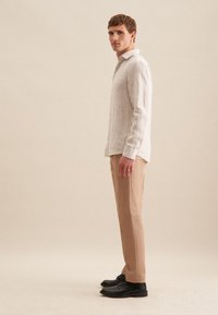 Gestreepte lange mouwen shirt in beige en wit, gecombineerd met beige broeken en zwarte schoenen, gefotografeerd in profiel tegen een neutrale achtergrond.