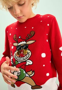 Rød strikket sweater med en tegneserie-type rensdyr iført solbriller, som spiller på en grøn guitar, omgivet af hvide snefnugsmønstre.