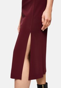 Burgunderfarbenes, figurbetontes Kleid mit einem seitlichen Schlitz, gefertigt aus glattem Stoff. Verfügt über einen konturierten Saum und einen sauberen Abschluss, der das Bein betont.