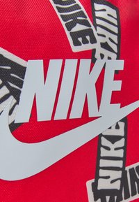 Tejido rojo de Nike con gran logo blanco y texto negro sobre acentos de patrón a rayas. El texto es atrevido y estilizado, mostrando la marca de manera prominente.