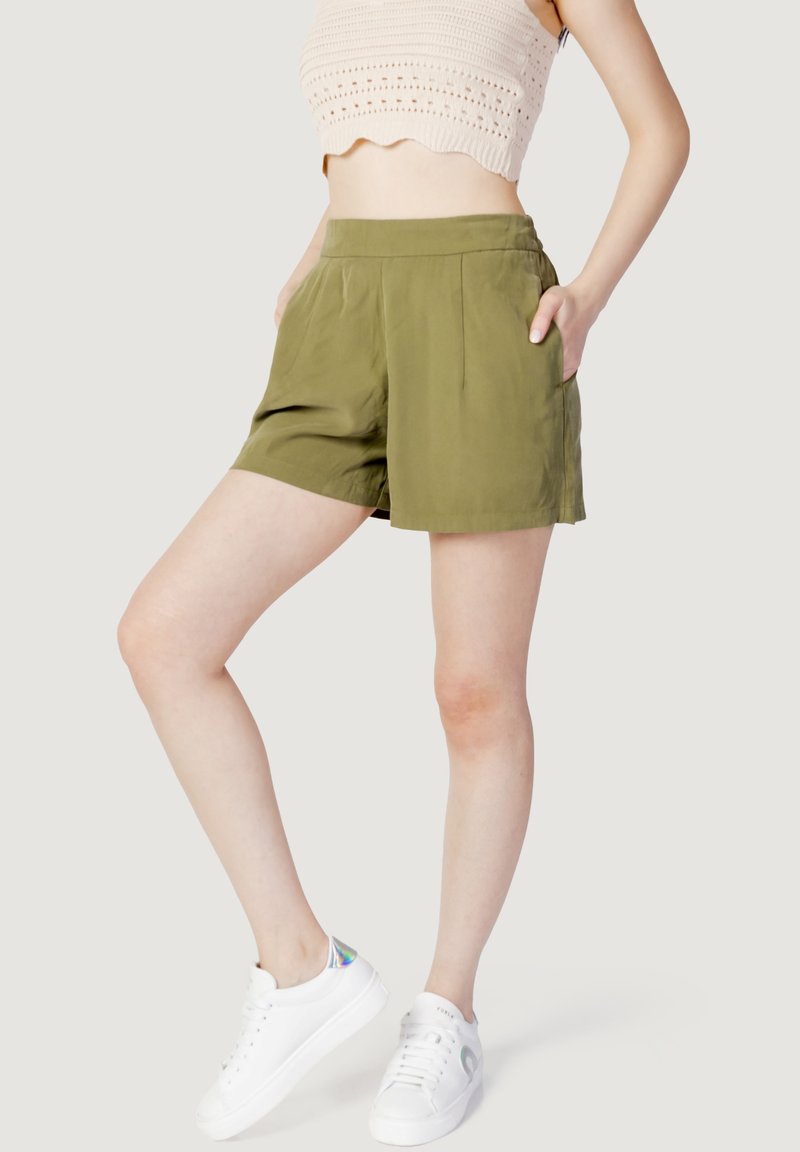Femme portant un short vert olive, un top court en maille beige et des baskets blanches, debout les mains dans les poches.