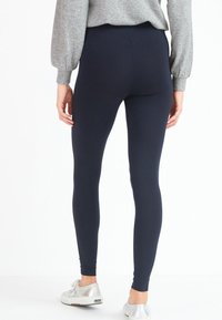 Marineblauwe leggings van rekbaar materiaal met een gladde textuur, gecombineerd met een grijze trui en zilveren sneakers, gezien vanuit de achterkant.