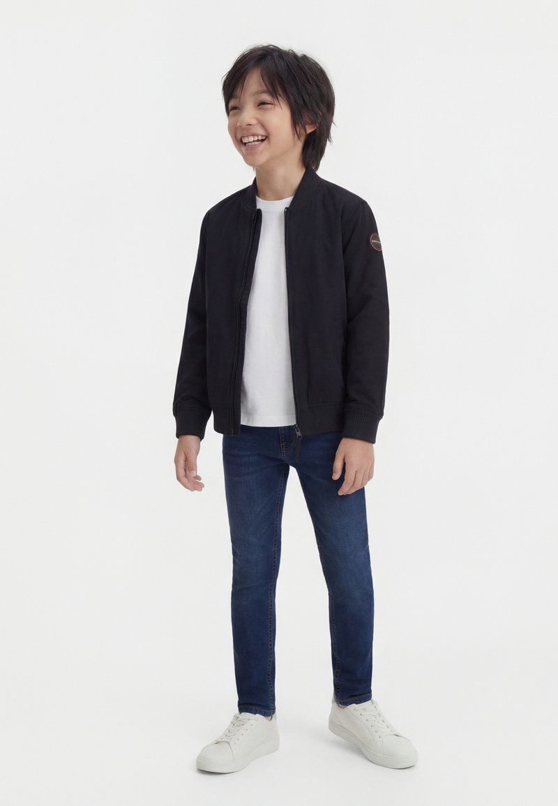 Χαμογελαστός αγόρι που φοράει μαύρο μπουφάν με φερμουάρ πάνω από ένα λευκό πουκάμισο, μπλε τζιν και λευκά αθλητικά παπούτσια, στέκεται μπροστά σε ένα απλό φόντο.