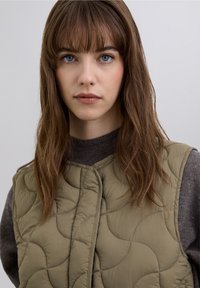 Olive green quilted vest with a rounded pattern, featuring a front snap closure. Underneath, a dark gray sweater is visible.