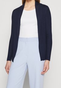 Námořnický žebrovaný cardigan s otevřeným předním dílem a dlouhými rukávy, nošený přes bílé tričko a světle modré kalhoty. Hladká textura, bez viditelných zapínání.