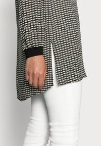 Haut long à manches noires avec un motif à fleurs orange et blanches ; possède des fentes sur les côtés et des poignets côtelés noirs, associé à un pantalon blanc.