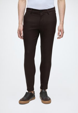 Homme portant un pantalon slim marron foncé, une chemise blanche et des baskets noires à semelles marron, debout devant un fond clair uni.