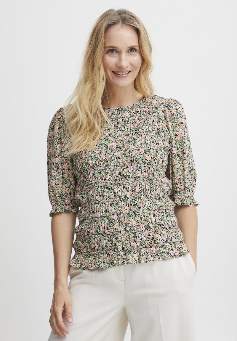 Blomsterbluse i grøn med pink og hvide mønstre. Har puffærmer og samlet tekstur i taljen. Parret med lyse bukser.