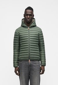 Vihreä toppatakki vaakasuoralla tikkauskuviolla, hupulla ja vetoketjulla. Varustettu oranssilla vetoketjun vedinnauhalla, yhdistetty tummiin farkkuihin.