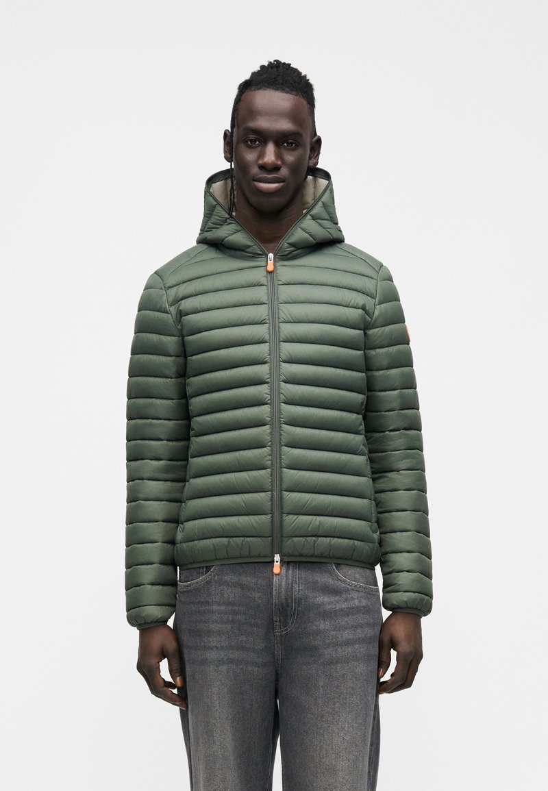 Vihreä toppatakki vaakasuoralla tikkauskuviolla, hupulla ja vetoketjulla. Varustettu oranssilla vetoketjun vedinnauhalla, yhdistetty tummiin farkkuihin.