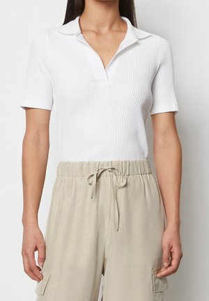 Person trägt ein weißes, geripptes kurzärmeliges Poloshirt, das in eine beige Hose mit Kordelzug und Seitentaschen gesteckt ist.