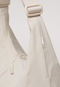 Sac en tissu beige au toucher lisse, doté d'une fermeture à glissière sur le dessus et d'une large bandoulière avec une boucle en plastique. Détail de petite étiquette visible.