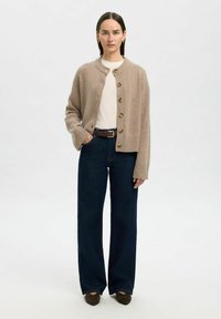 Cardigan en maille beige avec un col rond, fermeture à boutons et poignets côtelés, associé à un jean bleu foncé à jambes larges et des chaussures à talons marron.