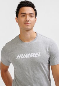 Camiseta gris de manga corta hecha de una tela suave, con un destacado logo blanco de "HUMMEL" en el pecho. Cuello redondo simple.