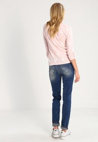 Ljusrosa långärmad topp med vita sjöstjärnemönster, i kombination med blå jeans. Modellen bär vita sneakers och visar upp uppvikta ärmar.