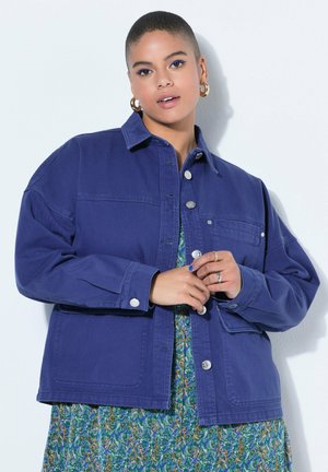 Młoda osoba z krótkimi włosami, ubrana w niebieską kurtkę na kwiecistej sukience, ozdobiona złotymi kolczykami-kołami i pierścionkami, na jednolitym tle.