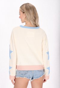 Felpa crema con accenti blu a stelle sulle maniche e un colletto azzurro chiaro, con orlo rosa e una toppa sul retro.