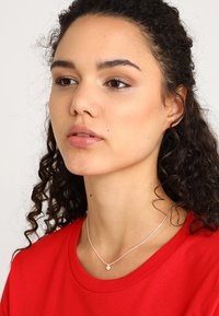 Silberne Halskette mit einem kleinen Herzanhänger, getragen über einem roten Shirt. Neutraler Hintergrund, der den Schmuck betont. Lockige Haare umrahmen das Gesicht.