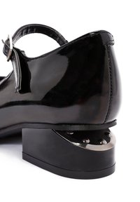 Zwarte lakleren schoen met een gesp, ronde neus en een onderscheidende metalen hak met klinknagels en een unieke vorm.