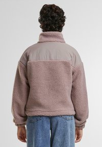 Osoba s kovrčavom tamnom kosom koja nosi mauve sherpa flis jaknu i plave traperice, okrenuta leđima prema jednostavnoj bijeloj pozadini.