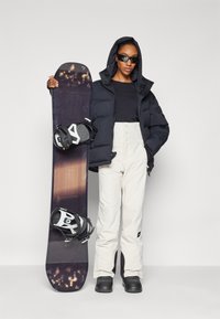 Schwarze Steppjacke, schwarzes Oberteil, weiße Schneehosen und schwarze Stiefel. Hält ein Snowboard mit schwarz-weißen Bindungen. Schwarze Sonnenbrille.