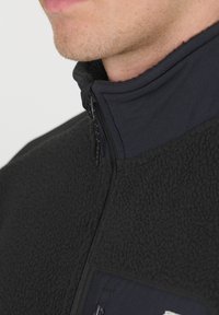 Veste en polaire noire avec un col montant, dotée d'un tissu texturé et d'une fermeture éclair à l'avant, agrémentée d'une poche unie bleu marine.