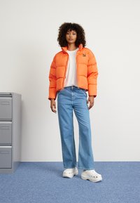 Giacca puffer arancione brillante, t-shirt bianca, jeans larghi blu e zoccoli bianchi spessi. L'ambientazione comprende un armadietto grigio e un tappeto blu.