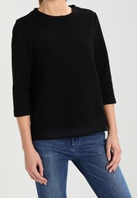 Sort tekstureret sweater med rund hals, trekvartærmer og lige kant, parret med blå jeans.