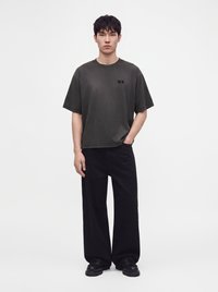 Νέος άνδρας που φοράει σκουρόγριφο oversized t-shirt, φαρδιά μαύρα παντελόνια και μαύρα chunky παπούτσια σε απλό λευκό φόντο.