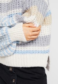 En person som bär en randig stickad tröja i blått, vitt, grått och beige, håller ärmen med sin hand; bär också svarta knäppbyxor.