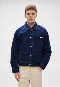 Mørkeblå denimjakke med krage, sølvknapper og hvite sømmer. Har et brystlomme med merkeetikett.