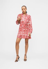 Γυναίκα με πλεξούδες, φοράει κόκκινο και λευκό μακρυμάνικο φόρεμα με σχέδιο, μαύρες ψηλοτάκουνες σανδάλες και κρατά καφέ τσάντα ώμου.