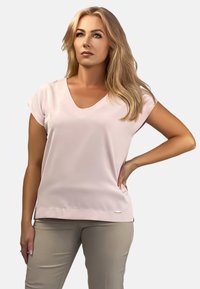 Jasnoróżowa bluzka z dekoltem w serek i krótkimi rękawami, o gładkiej fakturze, z subtelną metką marki na dole, zestawiona z jasnobeżowymi spodniami.
