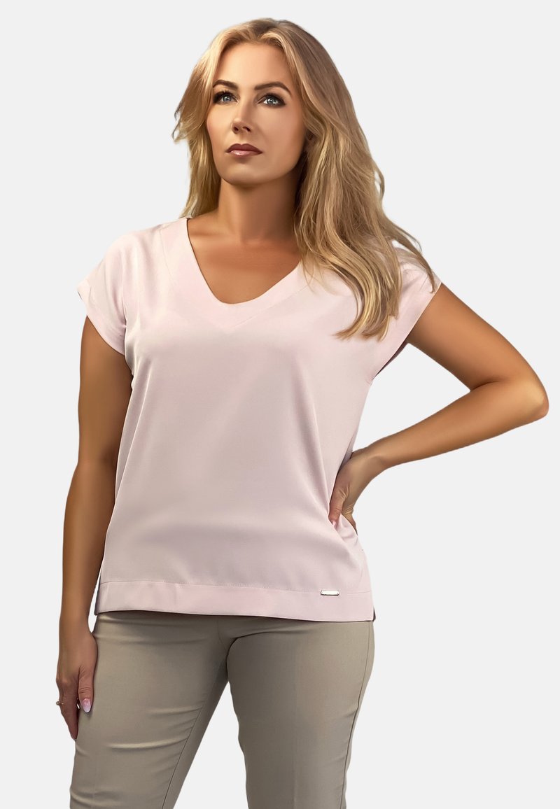 Jasnoróżowa bluzka z dekoltem w serek i krótkimi rękawami, o gładkiej fakturze, z subtelną metką marki na dole, zestawiona z jasnobeżowymi spodniami.