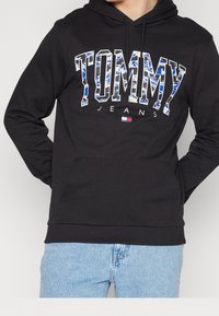 Sudadera negra de mezcla de algodón con un gran logo "TOMMY" en patrones azul y blanco, bolsillo tipo canguro y puños acanalados.