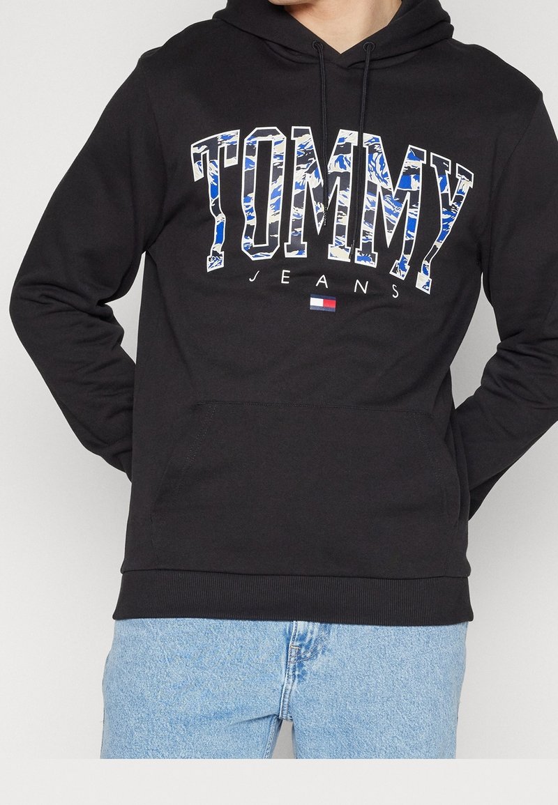 Sudadera negra de mezcla de algodón con un gran logo "TOMMY" en patrones azul y blanco, bolsillo tipo canguro y puños acanalados.