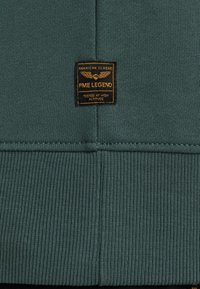 Grøn sweatshirt med ribbet struktur i kanten, med et sort og guld mærke der tydeligt viser "PME Legend".