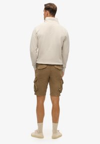 Pull en polaire beige avec un col haut, associé à un short cargo marron avec des poches latérales et un tissu texturé. Le modèle porte des baskets claires.