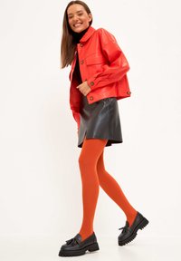 Giacca in ecopelle rossa, gonna in ecopelle nera, collant arancioni, mocassini neri con plateau e dettaglio a fiocco. Modella in piedi, sorridente.