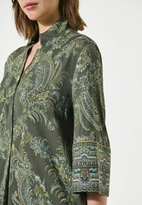 Groene blouse met een patroon van ingewikkelde bloemen- en paisleydesigns. De stof heeft een gladde textuur met een gedetailleerde manchetdetail.