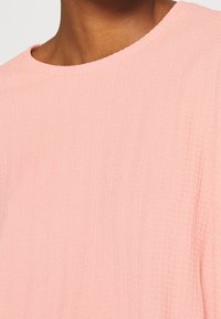 Vaaleanpunainen teksturoidun pinnan paita, jossa on pyöreä pääntie ja hienovaraiset pystyraitat, esittelee pehmeän, hieman ryppyisen kankaan yksityiskohtia.